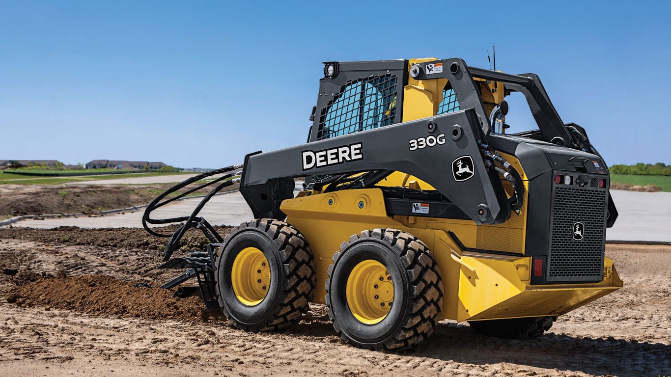 Une chargeuse à direction différentielle John Deere équipée de l’accessoire trancheuse en action sur un chantier.